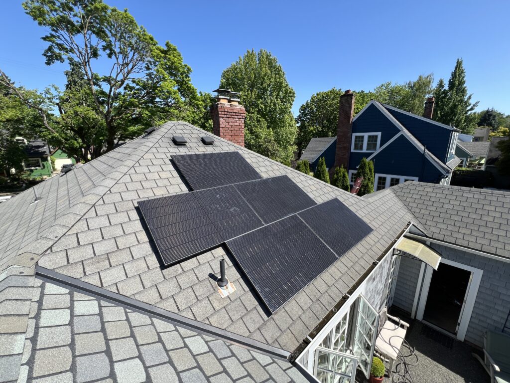 Black solar panels installed on a gray asphalt shingle roof of a residential home in a tree-lined neighborhood.