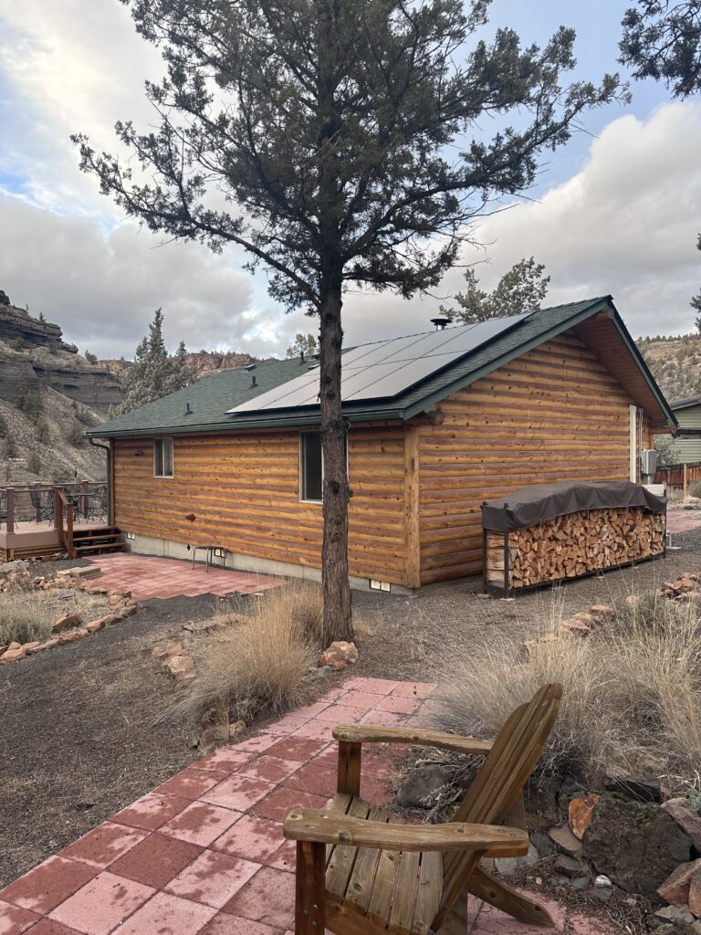 Black rooftop solar panels installed on a log cabin with a green asphalt shingle roof in a mountain landscape.