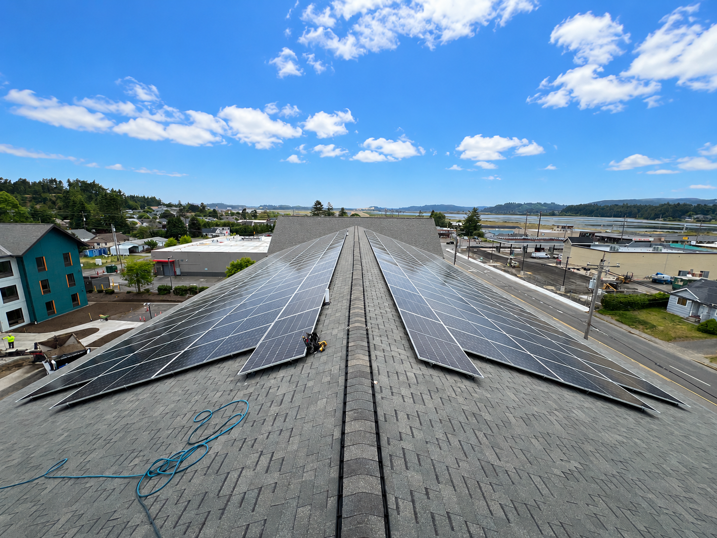 Large commercial rooftop solar array installed on a gray shingle roof beneath a bright blue sky, overlooking a coastal Oregon town.