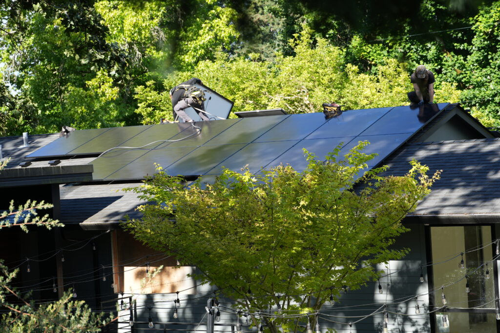 Solar installers mounting black solar panels on a residential roof surrounded by trees in Portland, Oregon.
