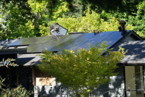 Solar installers mounting black solar panels on a residential roof surrounded by trees in Portland, Oregon.