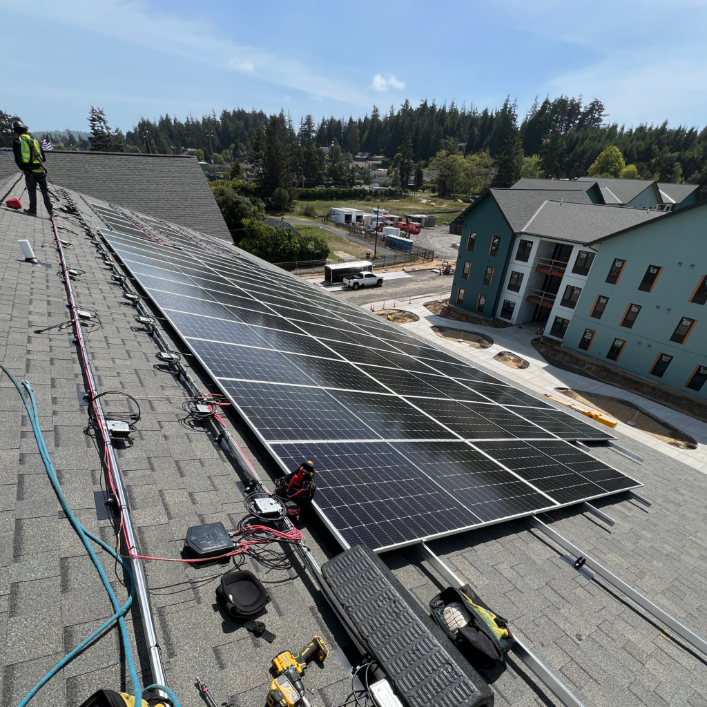 Rows of rooftop solar panels being installed on a multi-family housing building, with installers, electrical wiring, and a newly constructed apartment complex visible in the background under a clear blue sky.