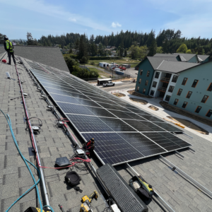 Rows of rooftop solar panels being installed on a multi-family housing building, with installers, electrical wiring, and a newly constructed apartment complex visible in the background under a clear blue sky.