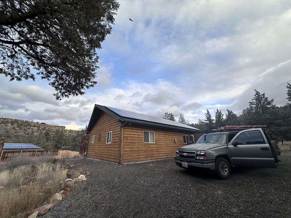 Solar panels installed on the metal roof of a rural log home surrounded by trees and open landscape.