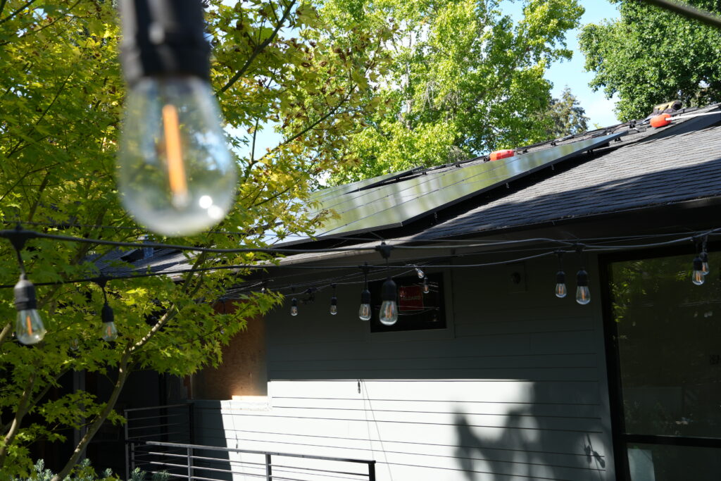 Solar panels installed on a residential roof surrounded by mature green trees and hanging patio lights.