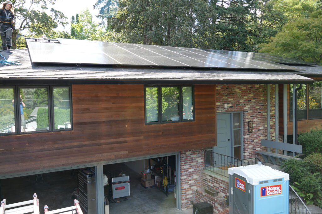 Rooftop solar panel array on a residential home surrounded by mature trees, with a solar installer working on the roof.